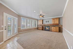 Unfurnished living room featuring ornamental molding, a fireplace with flush hearth, stone tile flooring, a ceiling fan, and light colored carpet