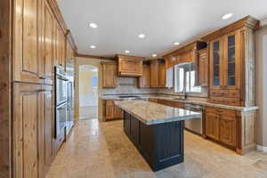 Kitchen with brown cabinetry, arched walkways, glass insert cabinets, stone tile flooring, and appliances with stainless steel finishes