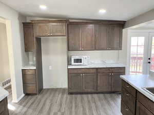 Kitchen with light countertops, white microwave, light wood-type flooring, dark brown cabinets, and recessed lighting