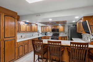 Kitchen with decorative backsplash, brown cabinets, light stone countertops, light tile patterned flooring, and a kitchen bar