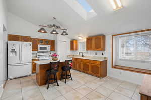 Kitchen featuring a skylight, lofted ceiling, white appliances, brown cabinetry, and light tile patterned flooring