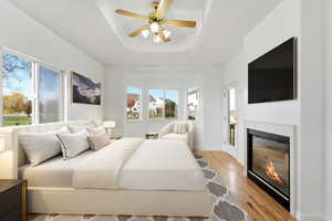 Virtually staged primary suite featuring a glass covered fireplace, light wood finished floors, a ceiling fan, and a tray ceiling