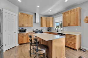 Kitchen featuring light brown cabinets, light wood-style flooring, recessed lighting, wall chimney range hood, and a breakfast bar