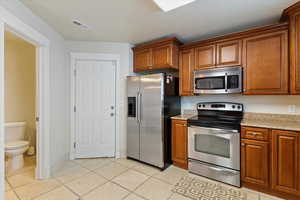 Kitchen with stainless steel appliances, brown cabinetry, light stone countertops, and light tile patterned flooring