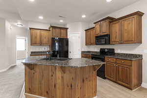 Kitchen featuring black appliances, a center island with sink, dark stone countertops, brown cabinets, and recessed lighting