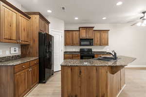 Kitchen featuring black appliances, brown cabinetry, dark stone countertops, an island with sink, and recessed lighting