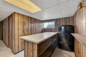 Interior space with wooden walls, a drop ceiling, freestanding refrigerator, light countertops, and light colored carpet