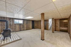 Basement with a wood stove, wooden walls, a paneled ceiling, light carpet, and stairway