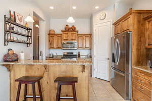 Kitchen with brown cabinetry, light stone counters, stainless steel appliances, a kitchen bar, and decorative light fixtures