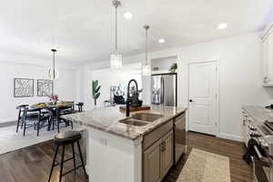 Kitchen featuring a kitchen island with sink, hanging light fixtures, stainless steel appliances, light stone countertops, and dark wood-type flooring