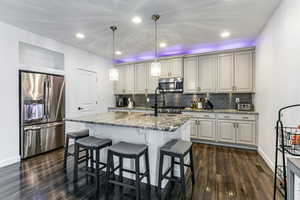 Kitchen featuring light stone counters, appliances with stainless steel finishes, hanging light fixtures, a breakfast bar, and an island with sink