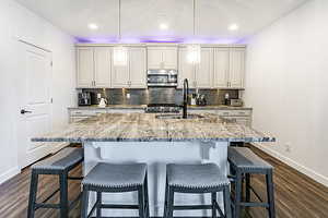 Kitchen featuring backsplash, light stone counters, dark wood-style floors, stainless steel appliances, and a kitchen bar