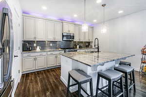 Kitchen with light stone countertops, stainless steel appliances, an island with sink, hanging light fixtures, and a kitchen bar