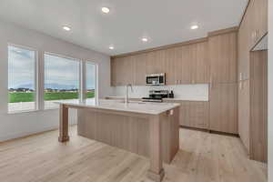Kitchen featuring modern cabinets, an island with sink, light stone counters, light brown cabinetry, and appliances with stainless steel finishes