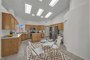 Kitchen featuring appliances with stainless steel finishes, brown cabinetry, high vaulted ceiling, a kitchen island, and light countertops