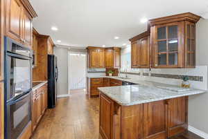Kitchen featuring brown cabinets, black appliances, tasteful backsplash, glass insert cabinets, and light stone countertops