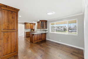 Kitchen featuring glass insert cabinets, a peninsula, brown cabinetry, light stone countertops, and dark wood-type flooring