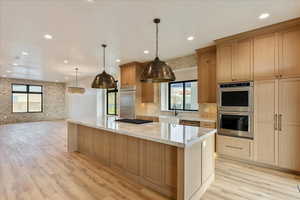 Kitchen with appliances with stainless steel finishes, light stone counters, light wood-style flooring, pendant lighting, and brick wall