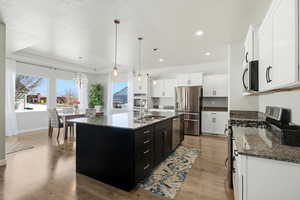 Kitchen featuring dark cabinetry, appliances with stainless steel finishes, hanging light fixtures, dark stone counters, and an island with sink