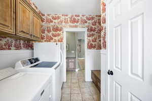 Washroom with wallpapered walls, wainscoting, cabinet space, washer and dryer, and light tile patterned flooring