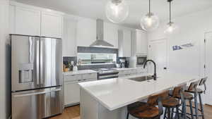 Kitchen featuring stainless steel appliances, white cabinets, wall chimney range hood, light wood finished floors, and a breakfast bar area