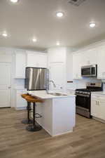 Kitchen featuring appliances with stainless steel finishes, a breakfast bar area, white cabinetry, an island with sink, and light wood finished floors