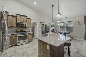 Kitchen featuring stainless steel appliances, light stone counters, high vaulted ceiling, decorative light fixtures, and a spacious island