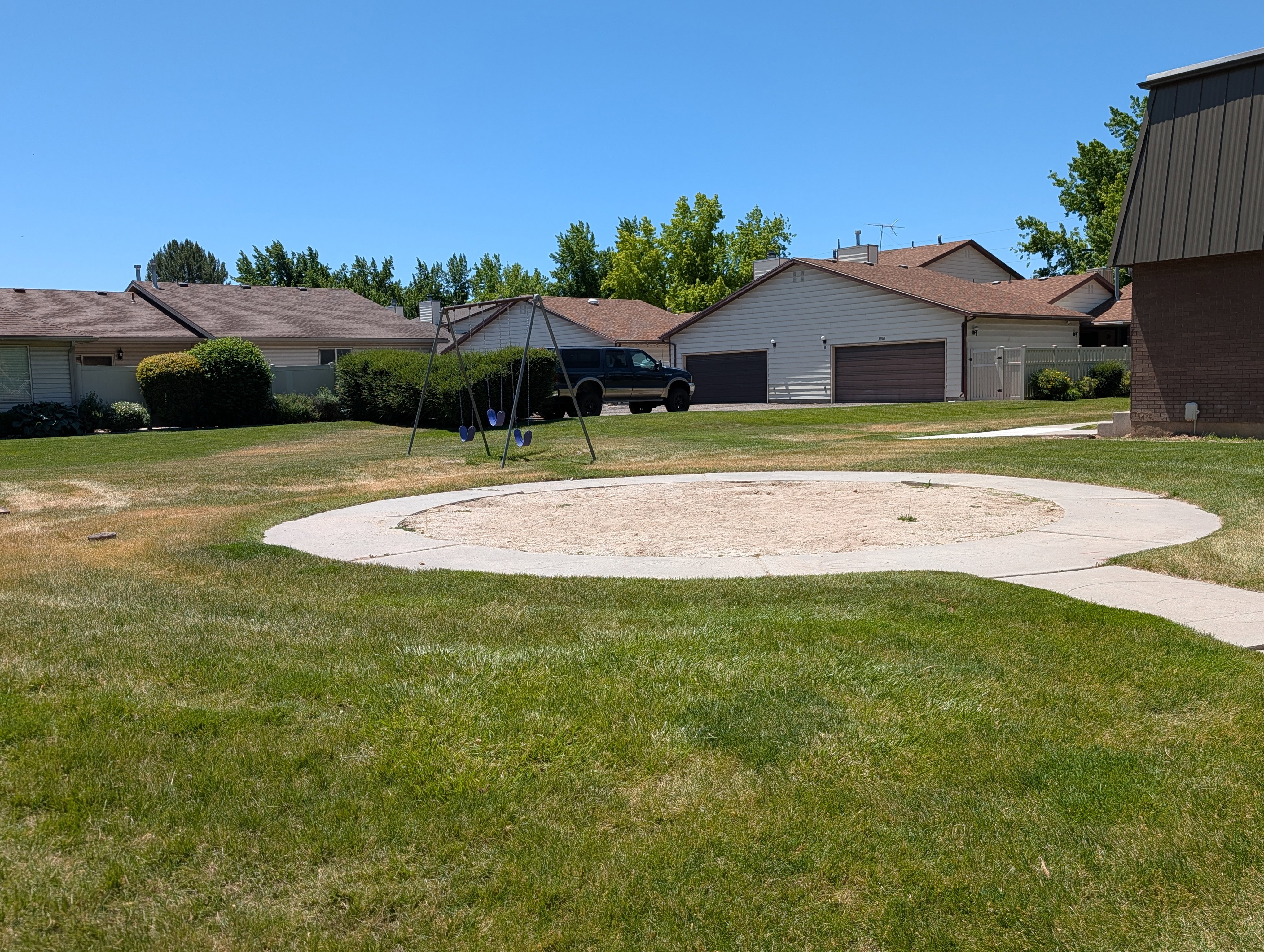 Community greenspace featuring a sand pit with a concrete border, a swing set, and a perimeter of mature trees