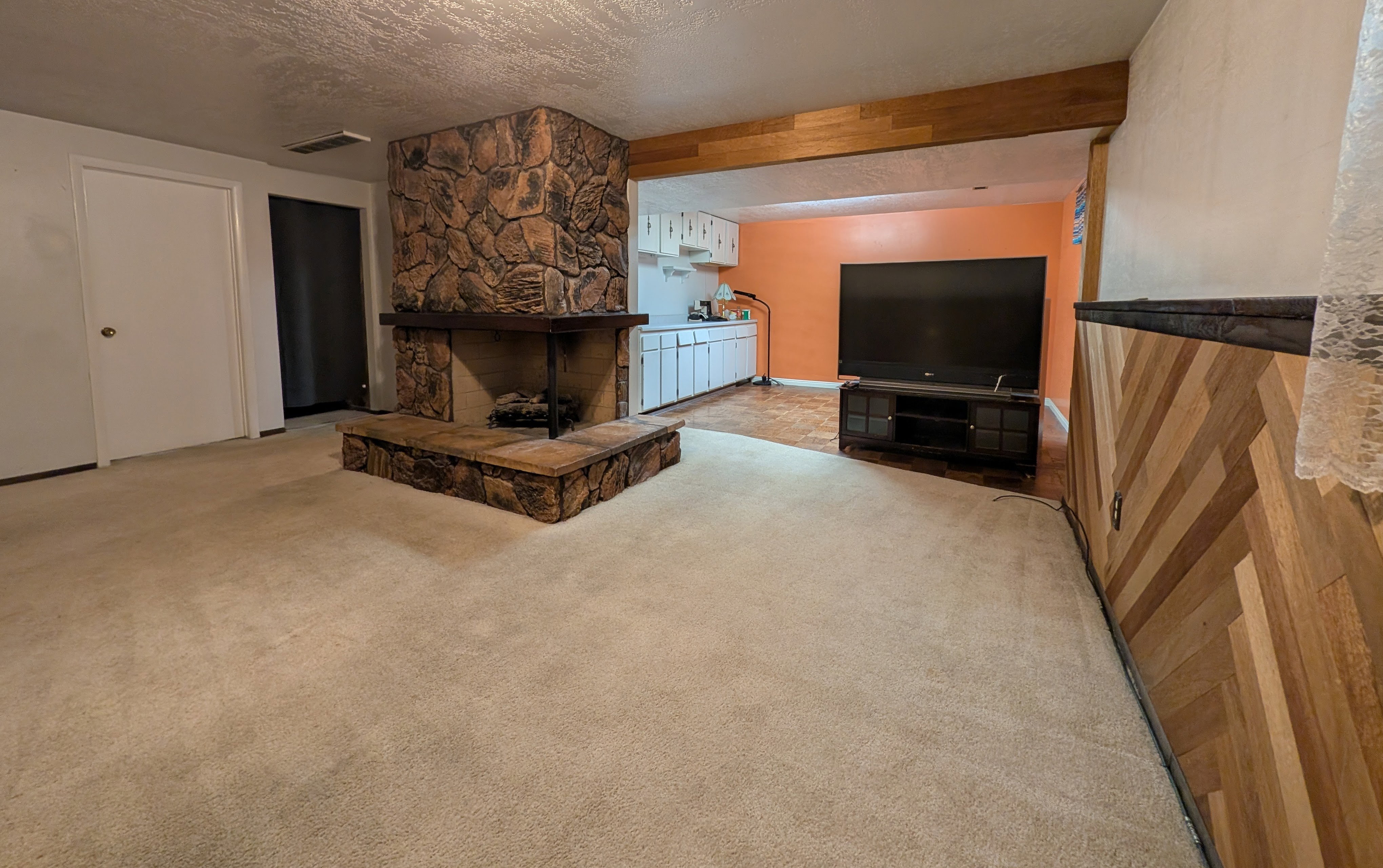 Living area featuring a stone-clad fireplace with a hearth, light-toned carpet, wood paneling with a diagonal pattern, and a wood beam ceiling accent