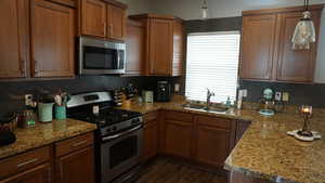Kitchen featuring stainless steel appliances, light stone countertops, decorative light fixtures, brown cabinetry, and dark wood-style floors