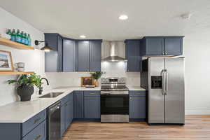 Kitchen featuring stainless steel appliances, open shelves, wall chimney range hood, light wood-type flooring, and light stone countertops