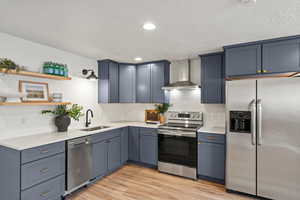 Kitchen featuring stainless steel appliances, wall chimney exhaust hood, recessed lighting, light wood-type flooring, and open shelves