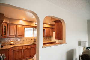 Kitchen featuring brown cabinets, light stone counters, a textured ceiling, glass insert cabinets, and tasteful backsplash