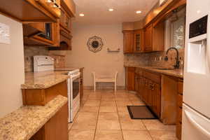 Kitchen with white appliances, glass insert cabinets, brown cabinets, light stone countertops, and light tile patterned floors