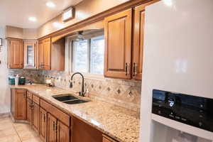 Kitchen featuring brown cabinets, white fridge with ice dispenser, light stone countertops, backsplash, and light tile patterned floors