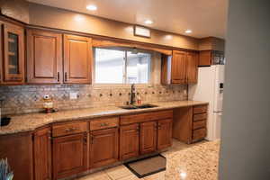 Kitchen featuring brown cabinetry, light stone counters, white fridge with ice dispenser, tasteful backsplash, and recessed lighting