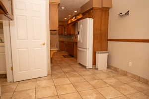 Kitchen with white refrigerator with ice dispenser, backsplash, brown cabinetry, glass insert cabinets, and light tile patterned flooring