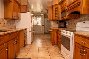 Kitchen featuring white appliances, brown cabinets, light stone countertops, and hanging light fixtures