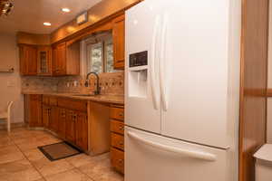 Kitchen featuring white refrigerator with ice dispenser, brown cabinetry, backsplash, light tile patterned floors, and light stone countertops