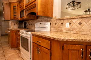 Kitchen featuring white range with electric stovetop, brown cabinetry, glass insert cabinets, light stone countertops, and light tile patterned flooring