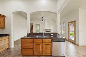 Kitchen with brown cabinetry, stainless steel dishwasher, ceiling fan, a tile fireplace, and dark stone counters