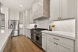 Kitchen featuring premium appliances, custom range hood, white cabinets, backsplash, and light wood-style flooring
