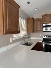 Kitchen with stainless steel appliances, light stone counters, brown cabinetry, and vaulted ceiling