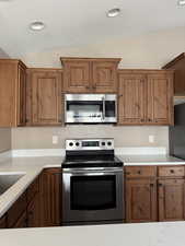 Kitchen with stainless steel appliances, brown cabinets, lofted ceiling, recessed lighting, and light stone counters