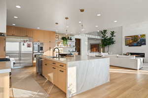 Kitchen with a large fireplace, light brown cabinetry, appliances with stainless steel finishes, hanging light fixtures, and an island with sink