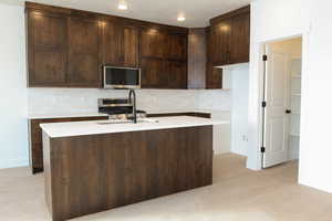 Kitchen featuring a kitchen island with sink, decorative backsplash, stainless steel appliances, dark wood finish cabinets, and light stone countertops