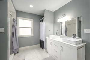 Full bath featuring double vanity, light marble finish flooring, and curtained shower