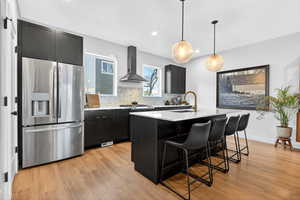 Kitchen with dark cabinets, stainless steel refrigerator with ice dispenser, decorative light fixtures, a breakfast bar, and wall chimney range hood