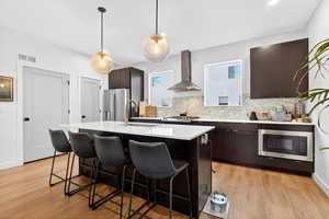 Kitchen with dark brown cabinets, wall chimney range hood, appliances with stainless steel finishes, a center island with sink, and a breakfast bar