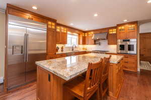 Kitchen featuring brown cabinetry, glass insert cabinets, appliances with stainless steel finishes, dark wood-style flooring, and recessed lighting